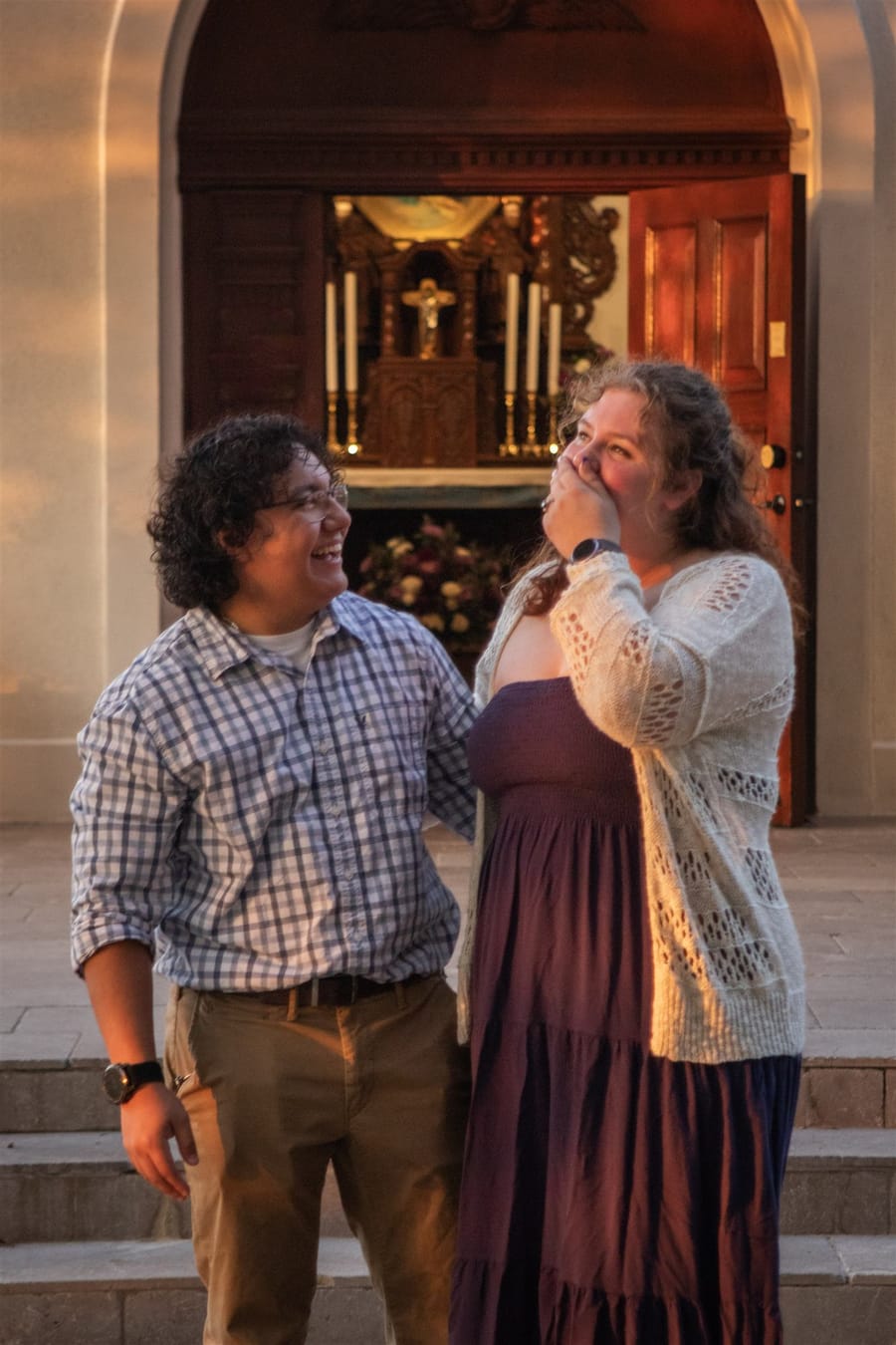 Salvador and Isabel laughing together in a chapel doorway at golden hour, with a gilded altar and lit candles behind them.
