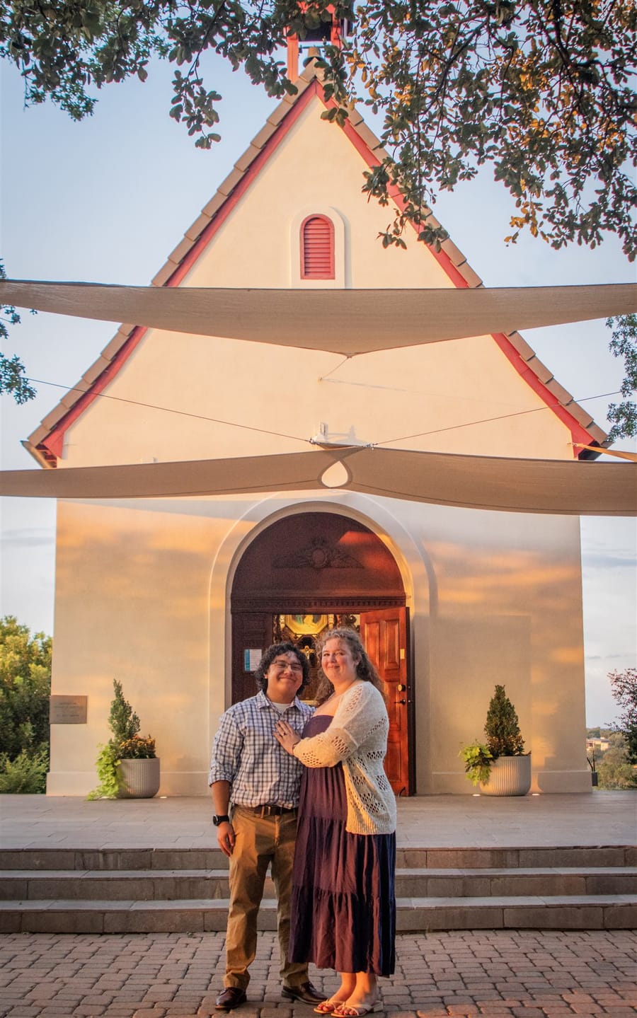 Salvador and Isabel standing together in front of a stucco chapel with a bell gable, at sunset.