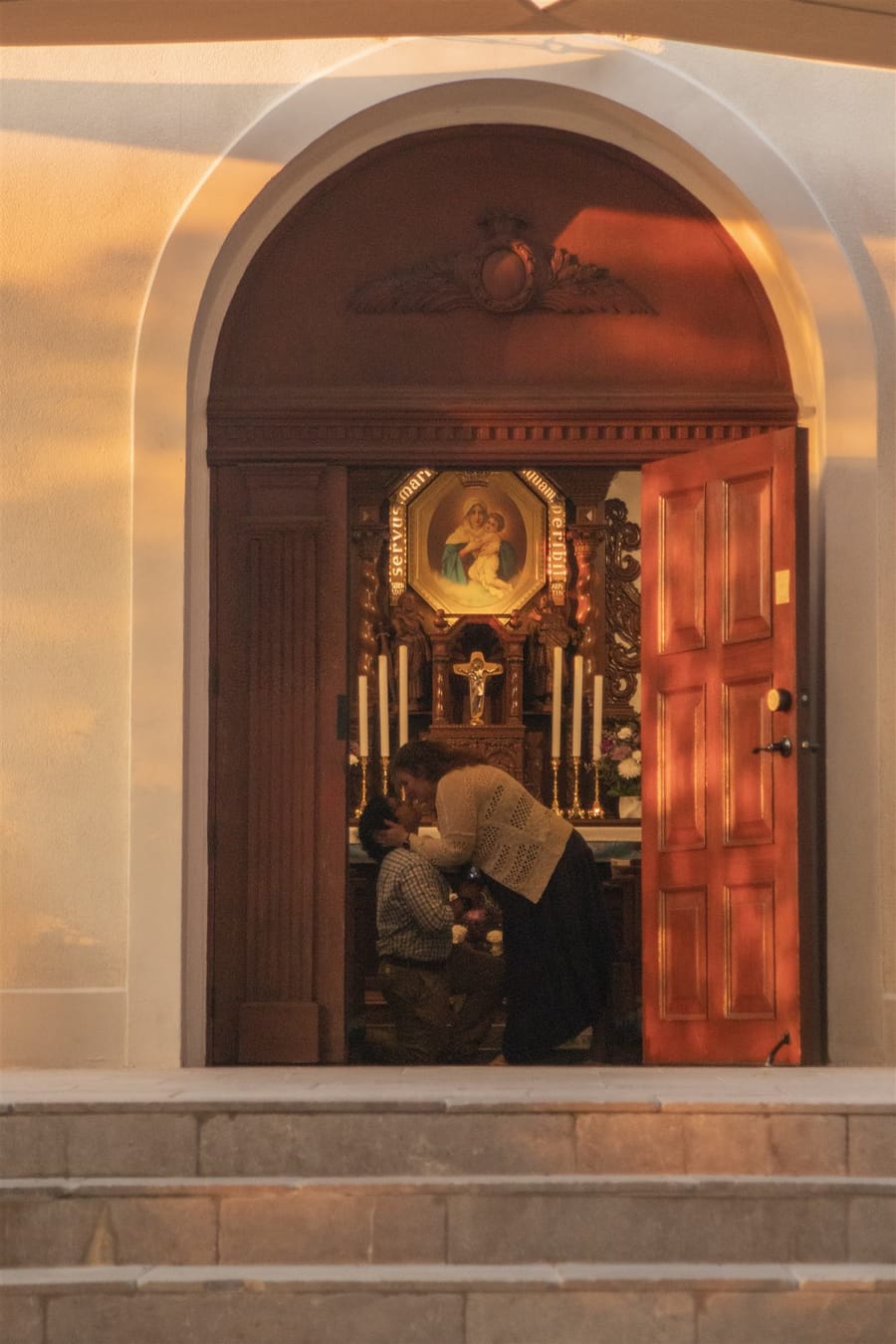 Salvador kneeling as he proposes to Isabel at the altar, candlelight and a gilded Marian icon behind them.